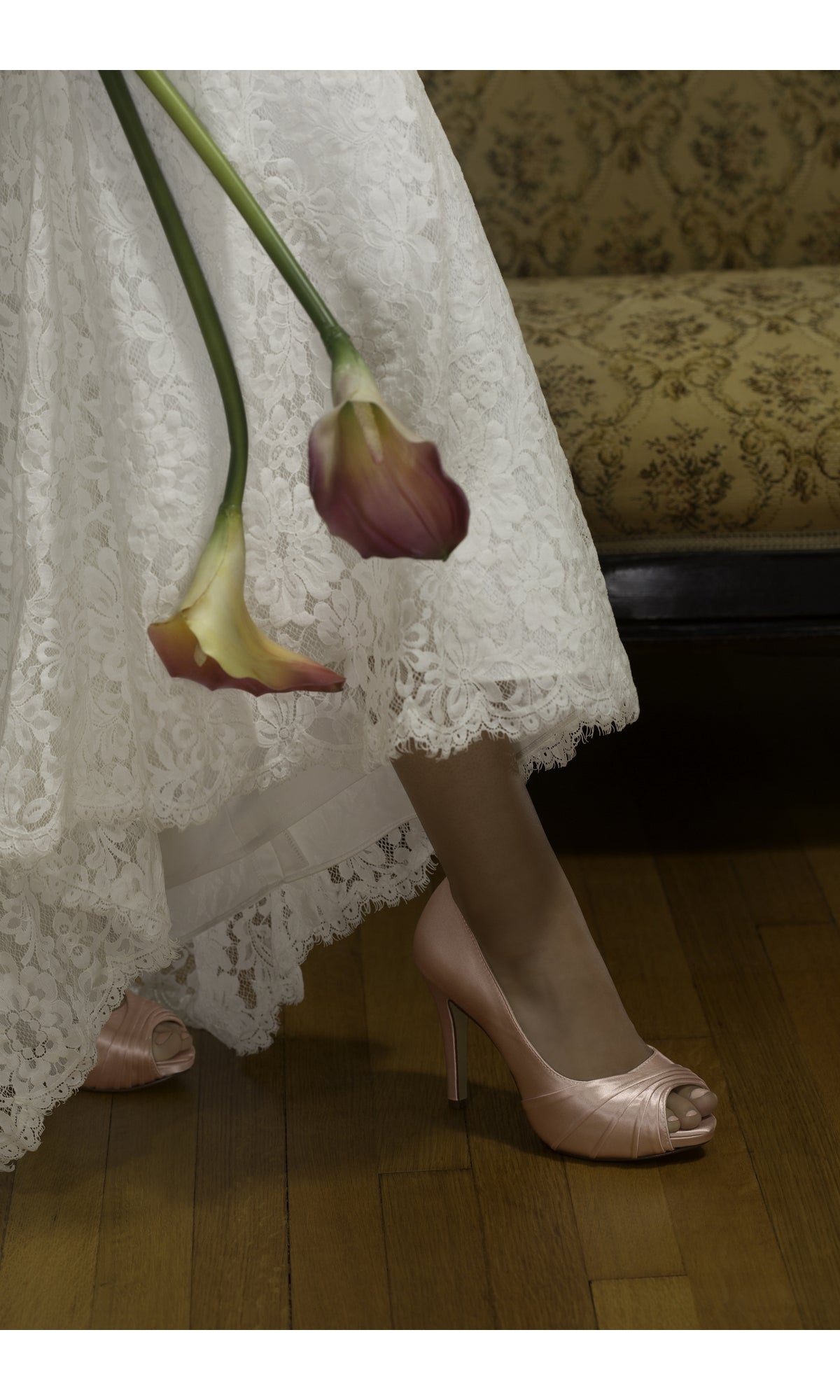 A woman in a white lace dress holds two calla lilies and wears Bea White Peep Toe Pump Prom Shoes by Dyeables 51716, standing on a wooden floor near an upholstered bench.